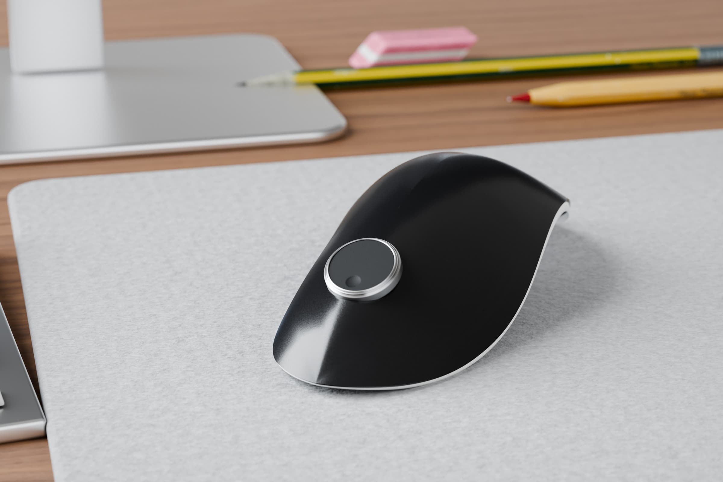 Horizon Model 11, a low semi-gloss black mouse with a brushed aluminium puck ring and a matte face, close up on a light felt desk mat in daylight, with a monitor stand, pencils and the edge of a keyboard behind.